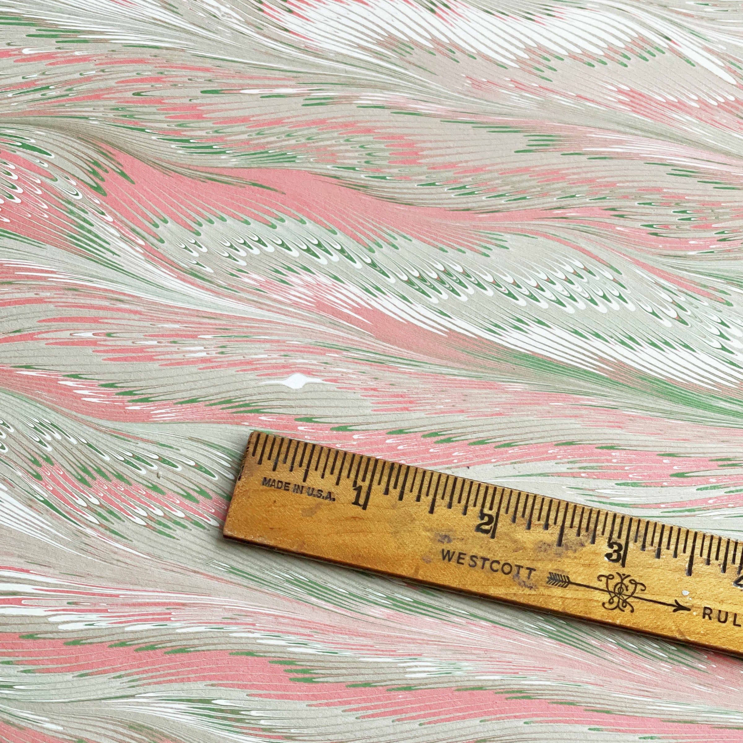 Marbled paper with a feathered pattern in pink and green with a wooden ruler for scale.